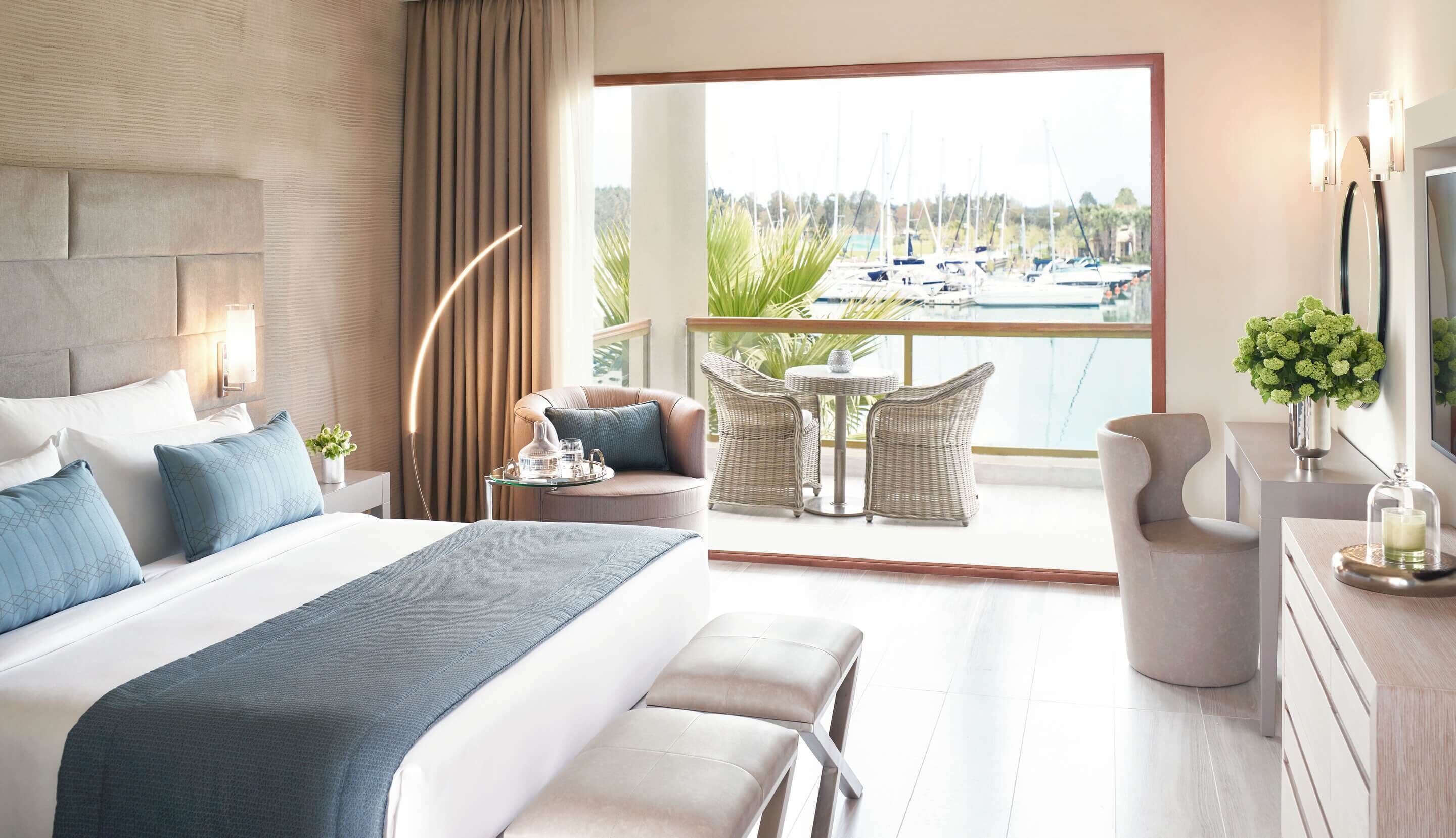 A modern hotel room with a large bed, a balcony with a view of boats, wicker furniture, and a vase of flowers.