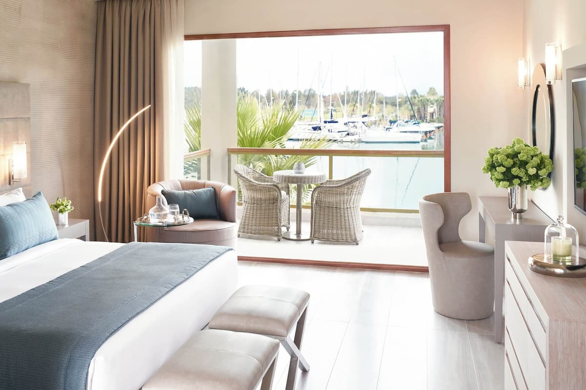 A modern hotel room with a large bed, a balcony with a view of boats, wicker furniture, and a vase of flowers.