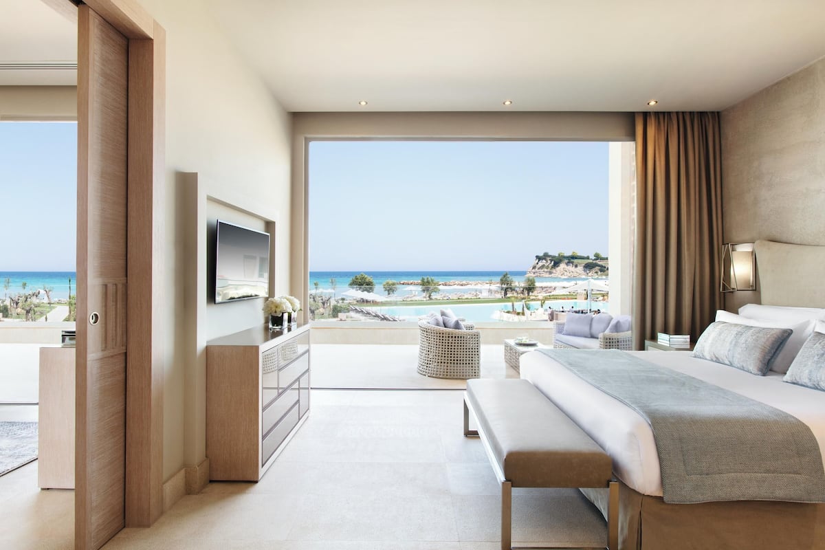 A modern hotel room with a large balcony, a bed, a TV, and a view of the beach and ocean.