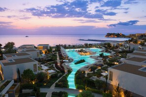 A resort with a central swimming pool, connected by walkways, surrounded by modern buildings and a view of the sea at dusk. - Sani Dunes (Kassandra)