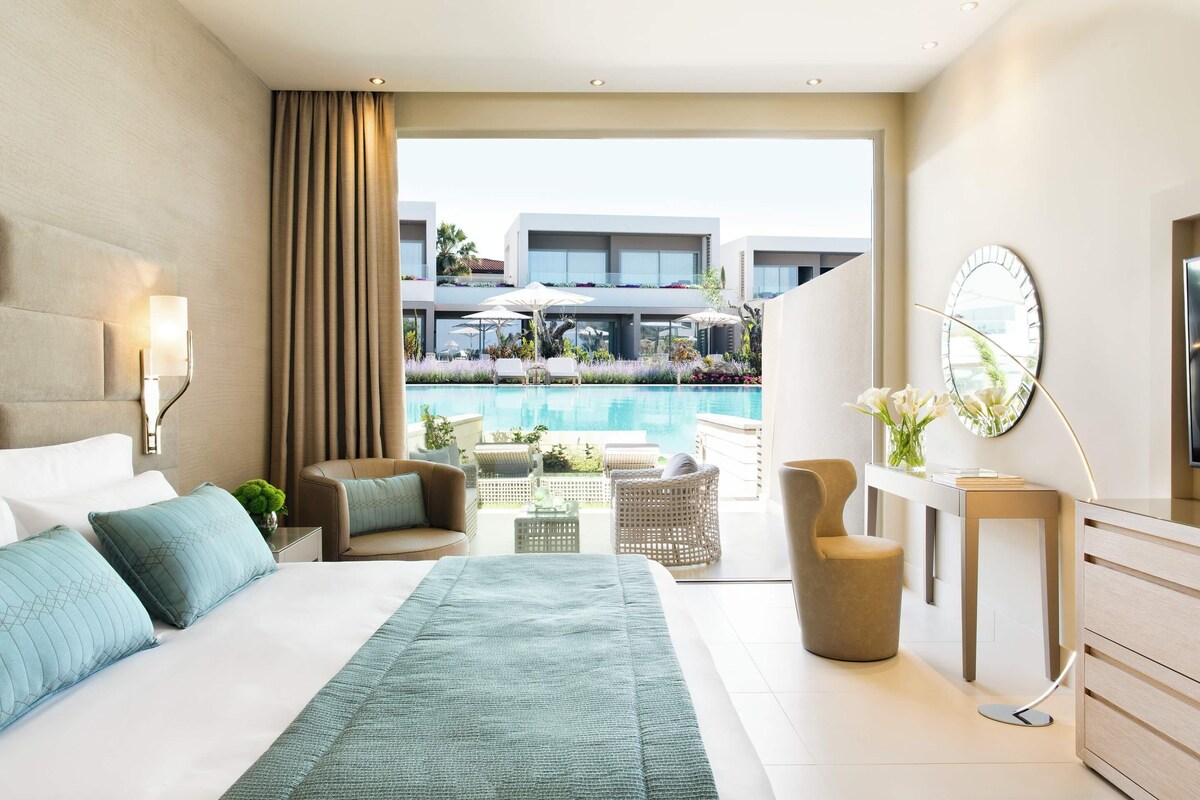 A modern hotel room with a large bed, a view of a pool, and a balcony with outdoor furniture.