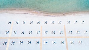 Eine Vogelperspektive zeigt einen Strand mit liegenden Liegestühlen und Sonnenschirmen, einem klaren, blauen Meer und einem sandigen Ufer.