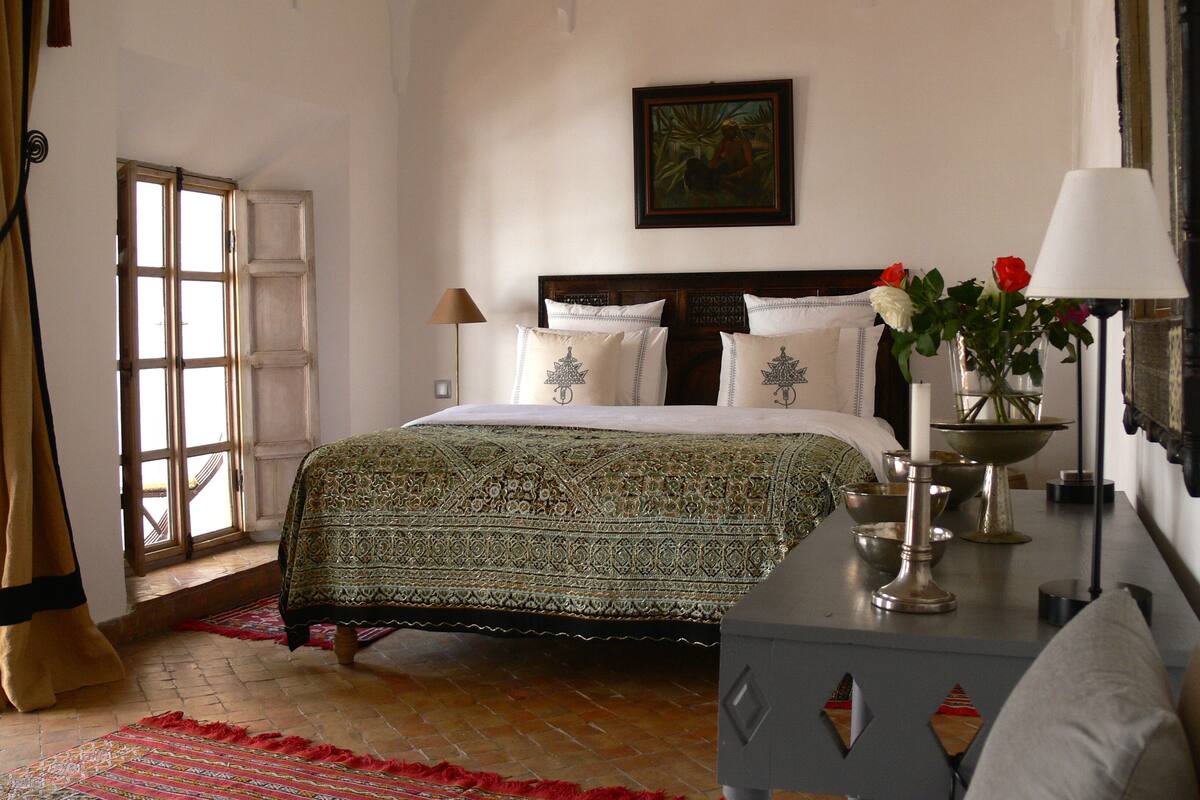 A bedroom with a large bed, a nightstand, a window, and a painting on the wall.