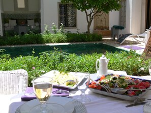An outdoor dining area with a table set for two, a white teapot, a glass of white wine, and a plate of salad. - Riad El Mezouar (Marrakech)