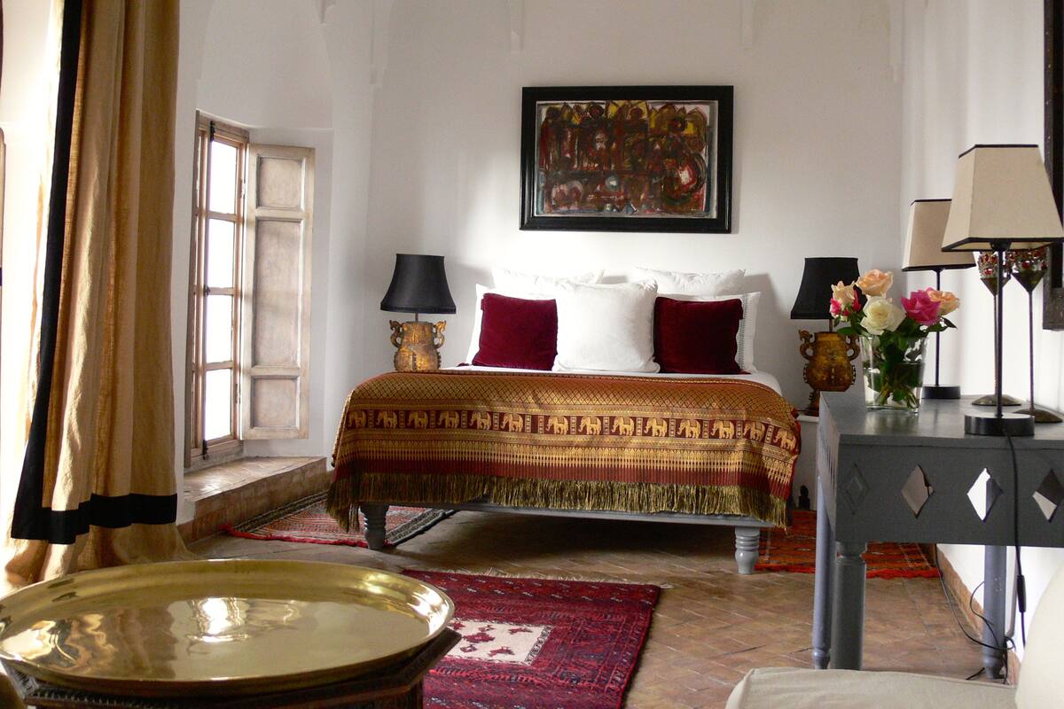 A hotel room with a bed, a painting on the wall, two lamps, a vase with flowers, and a large brass tray.