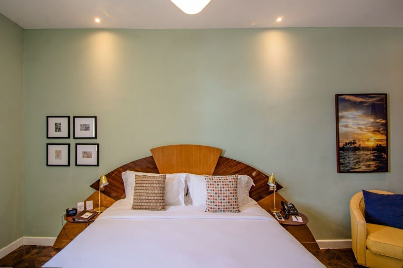 Habitación de hotel con una cama grande, cabecera de madera, mesita de noche con lámpara, una silla y un cuadro en la pared.