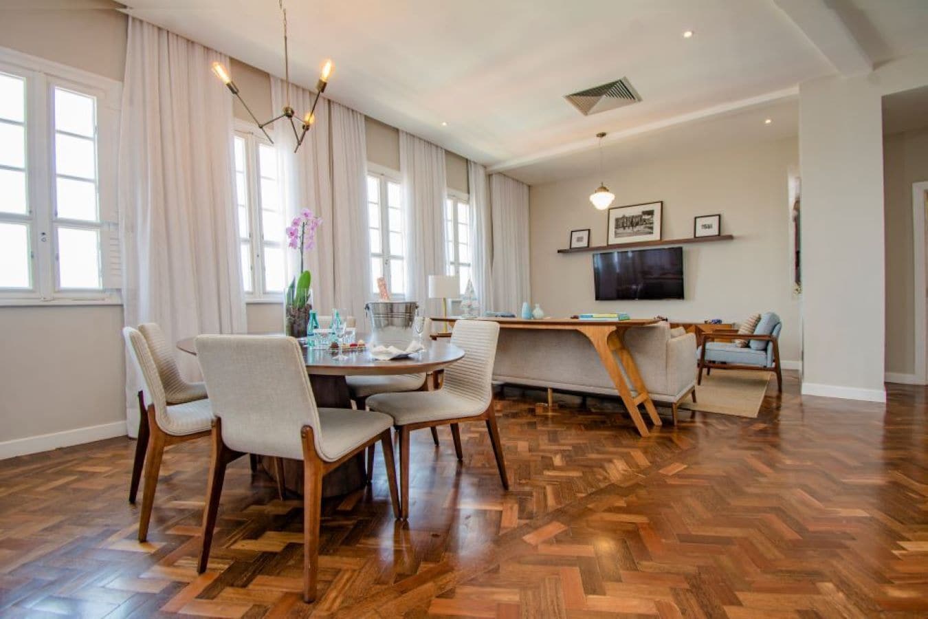 Amplia sala de estar con suelo de madera en patrón de espiga, una mesa redonda de comedor con sillas, un sofá y un televisor montado en la pared.