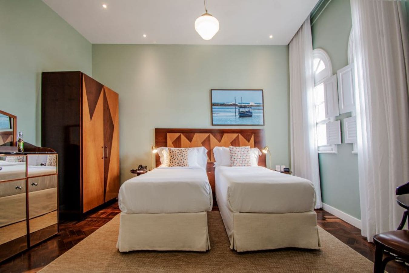 A hotel room with two beds, a wooden wardrobe, a desk with a chair, and a large window with curtains.
