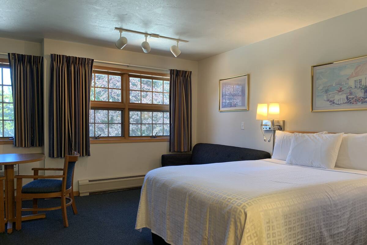 Ein Hotelzimmer mit einem großen Bett, einem Schreibtisch mit Stuhl, einem Fenster mit Vorhängen, einer Wandlampe und einem gerahmten Bild.