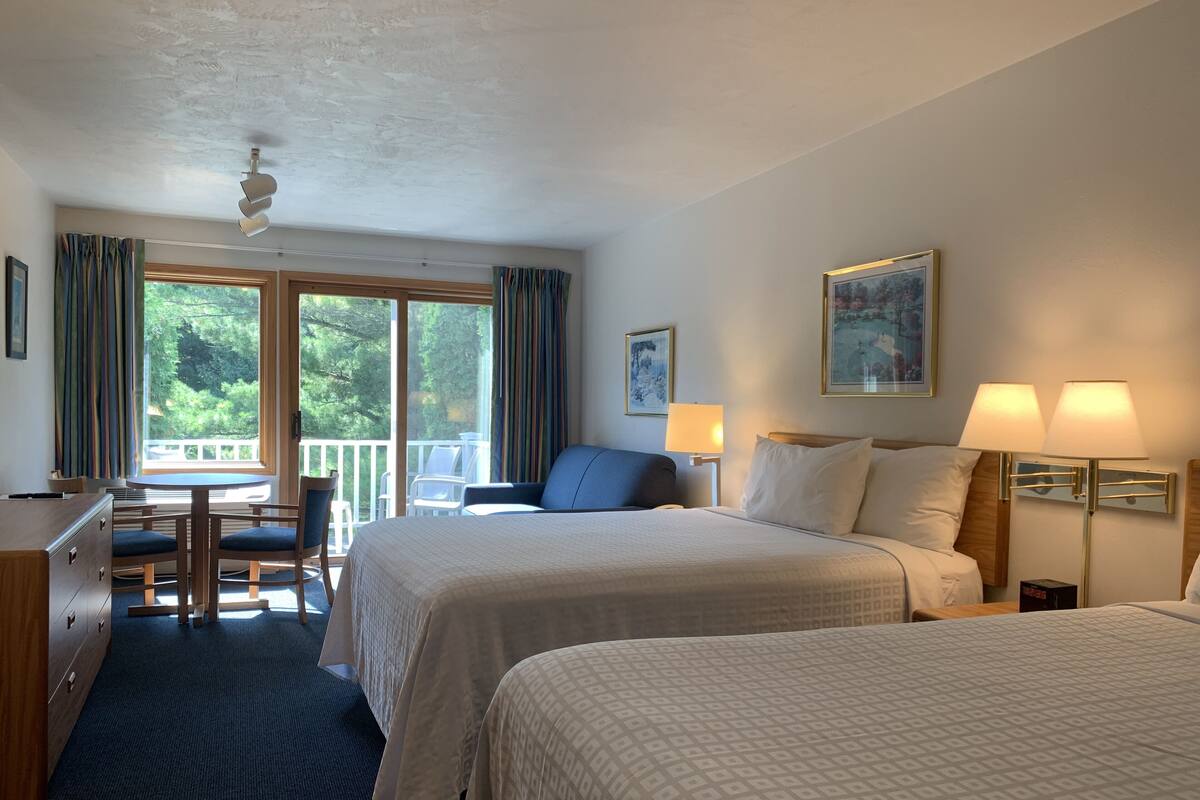 Ein Hotelzimmer mit zwei Betten, einem Schreibtisch, einem Stuhl, einem Balkon mit Blick auf Bäume und einem Deckenventilator.