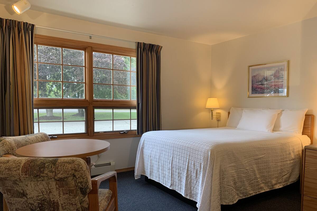 Ein Hotelzimmer mit einem Bett, einem Schreibtisch, einem Stuhl, einem Fenster mit Vorhängen, einer Lampe und einem Bild an der Wand.