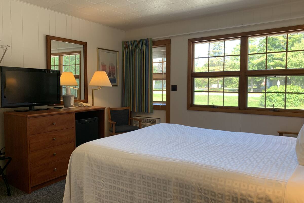 Ein Hotelzimmer mit Bett, Schreibtisch mit Fernseher, Stuhl, Kommode und großen Fenstern mit Blick auf Grünflächen.