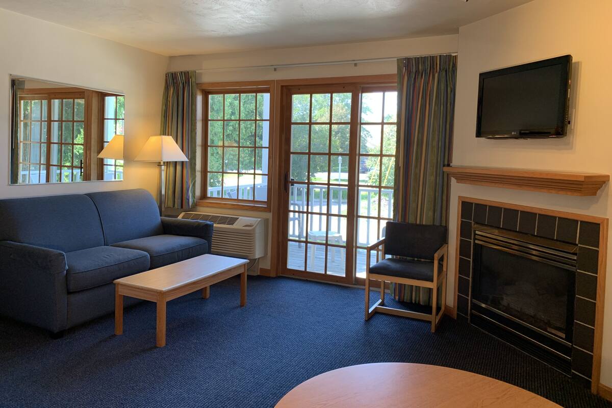 Ein Hotelzimmer mit einer Couch, einem Sessel, einem kleinen Tisch, einem Kamin, einem an der Wand befestigten Fernseher und großen Fenstern mit Vorhängen.
