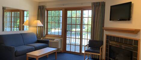 A hotel room with a sofa, a chair, a small table, a fireplace, a TV mounted on the wall, and large windows with curtains.