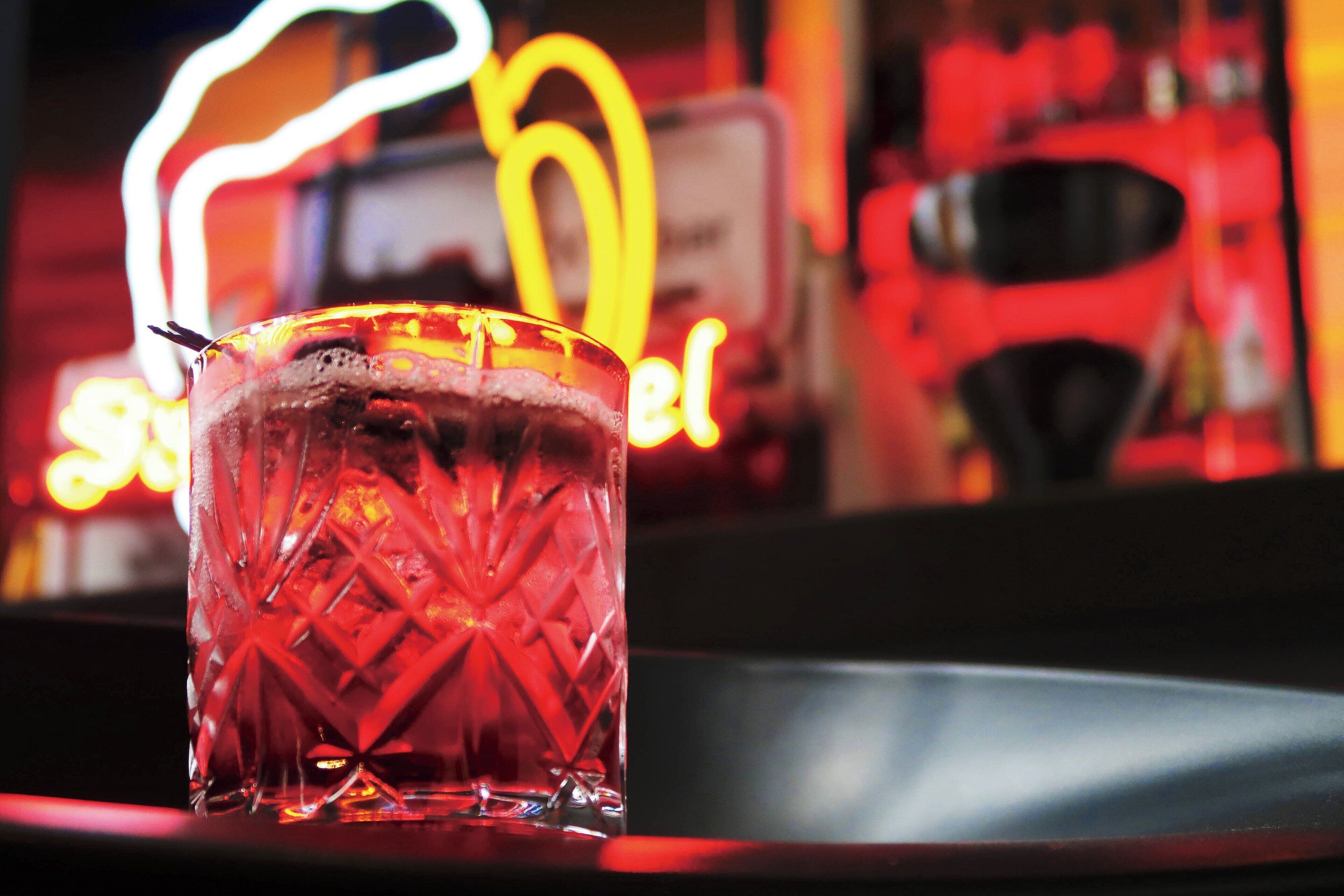 A glass with a textured pattern, placed on a surface with a blurred background featuring a neon sign.