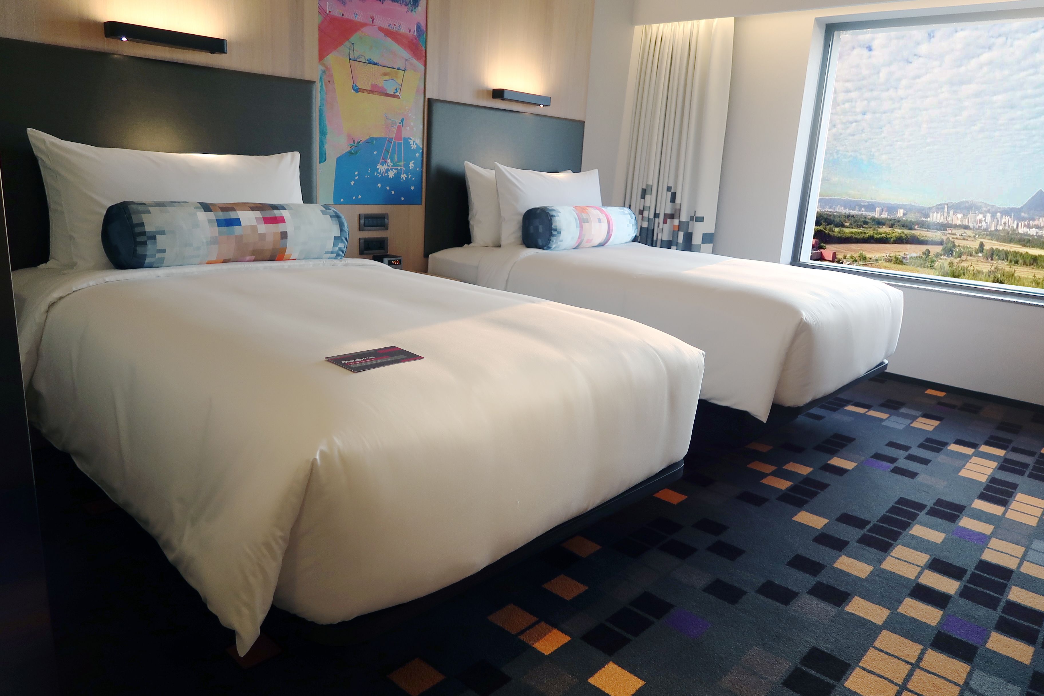 A hotel room with two beds, a large window with a city view, and a colorful abstract painting on the wall.