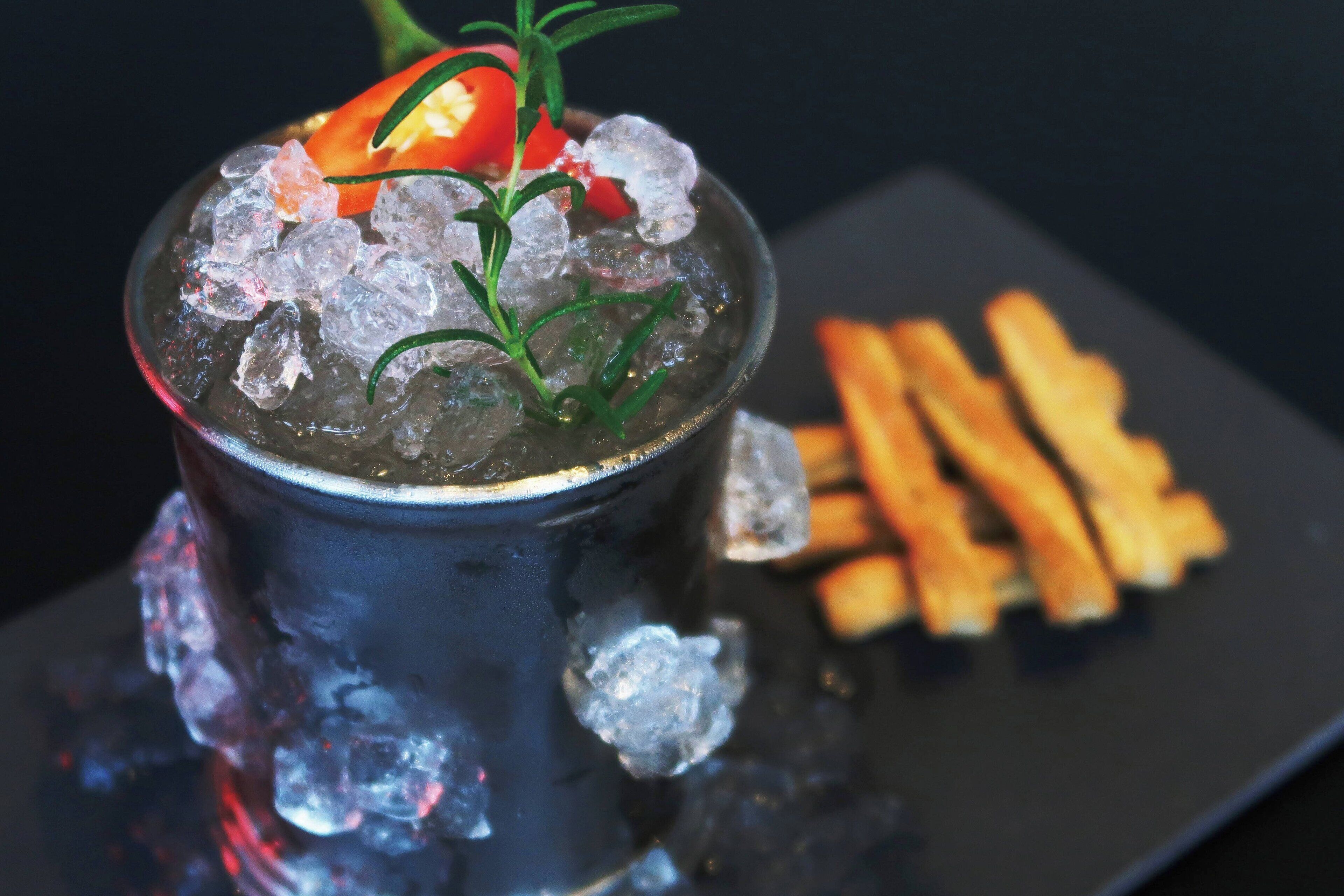 A cocktail in a glass with ice cubes and a sprig of rosemary, accompanied by a plate of fries.