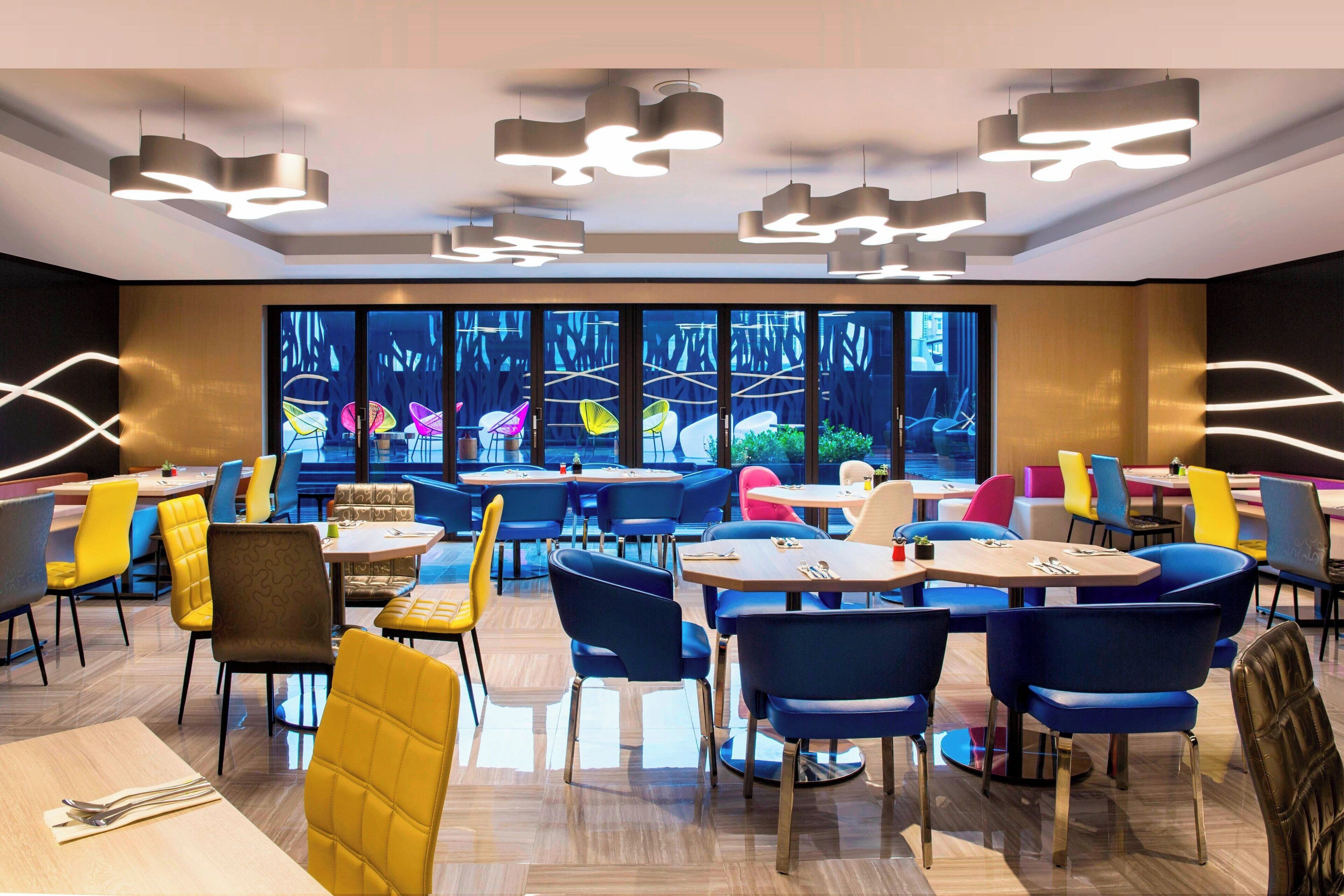 A modern dining area with multiple tables and chairs, large windows, and contemporary lighting fixtures.