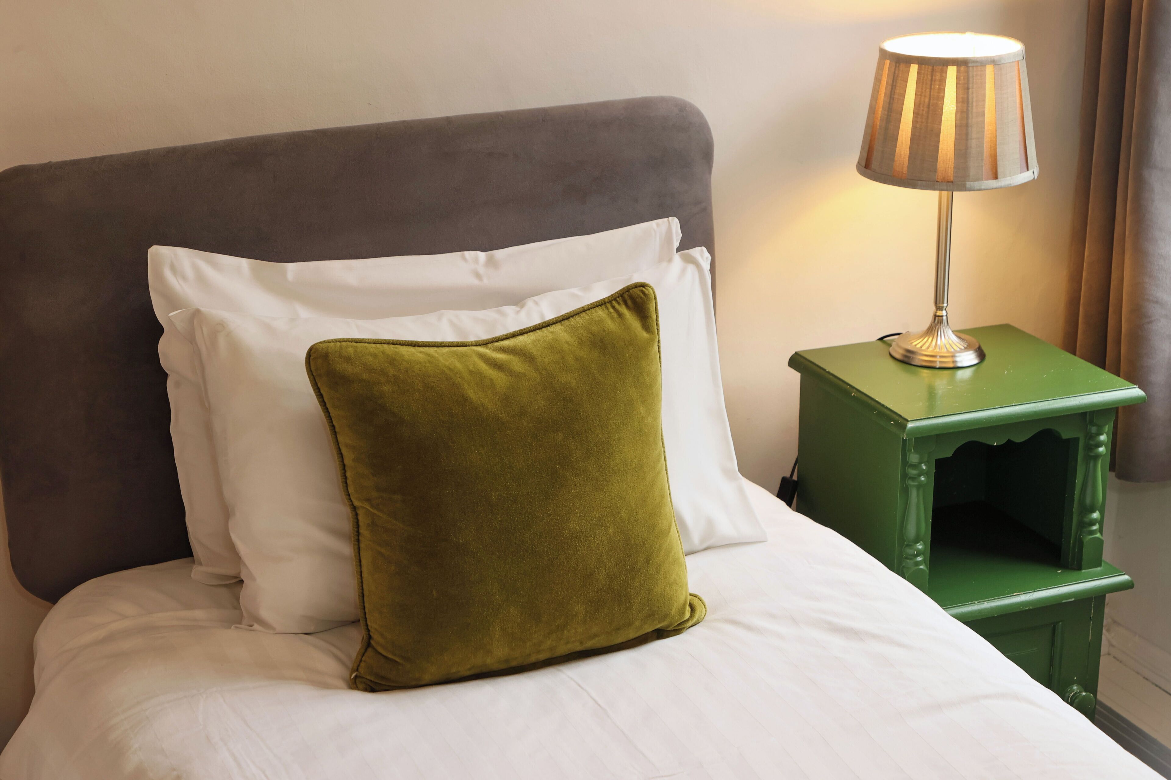 A bed with white bedding and a green pillow, a green bedside table with a lamp, and a grey headboard.