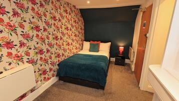 Een slaapkamer met bloemenbehang, een bed met een blauwgroene deken en een nachtkastje met een rode lamp.