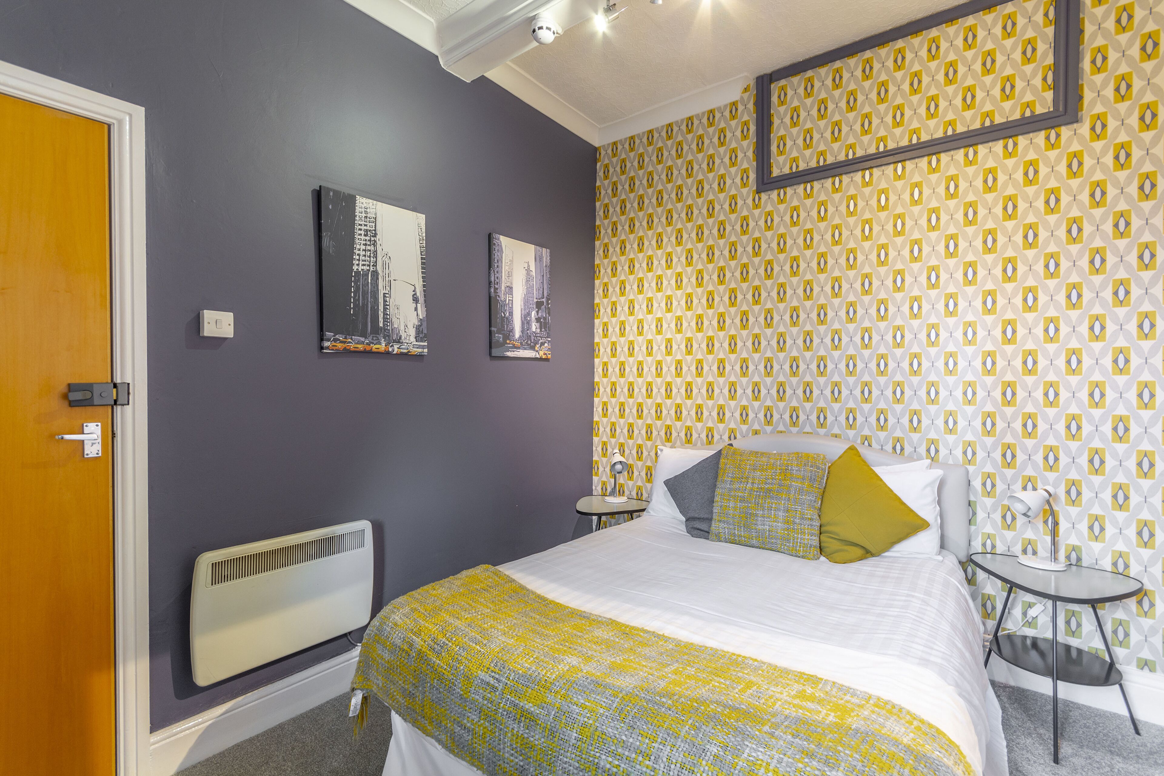 A bedroom with a bed, a radiator, two paintings, a patterned wallpaper, and a small table with a lamp.