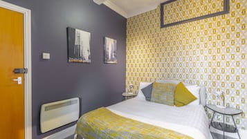 Een slaapkamer met een bed, een radiator, twee schilderijen, behang met een patroon en een klein tafeltje met een lamp.