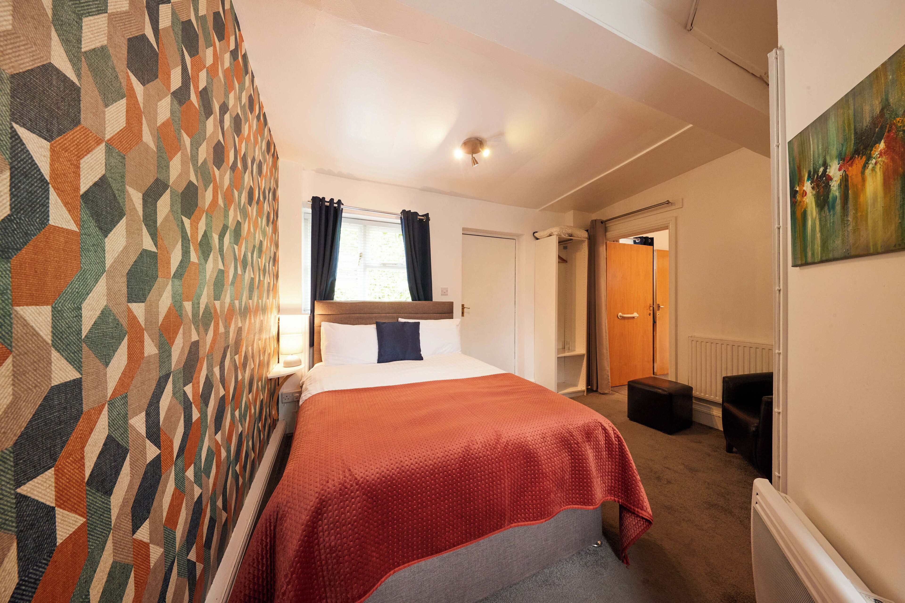 A hotel room with a patterned wall, a bed with a red blanket, a window with curtains, a door, and a radiator.