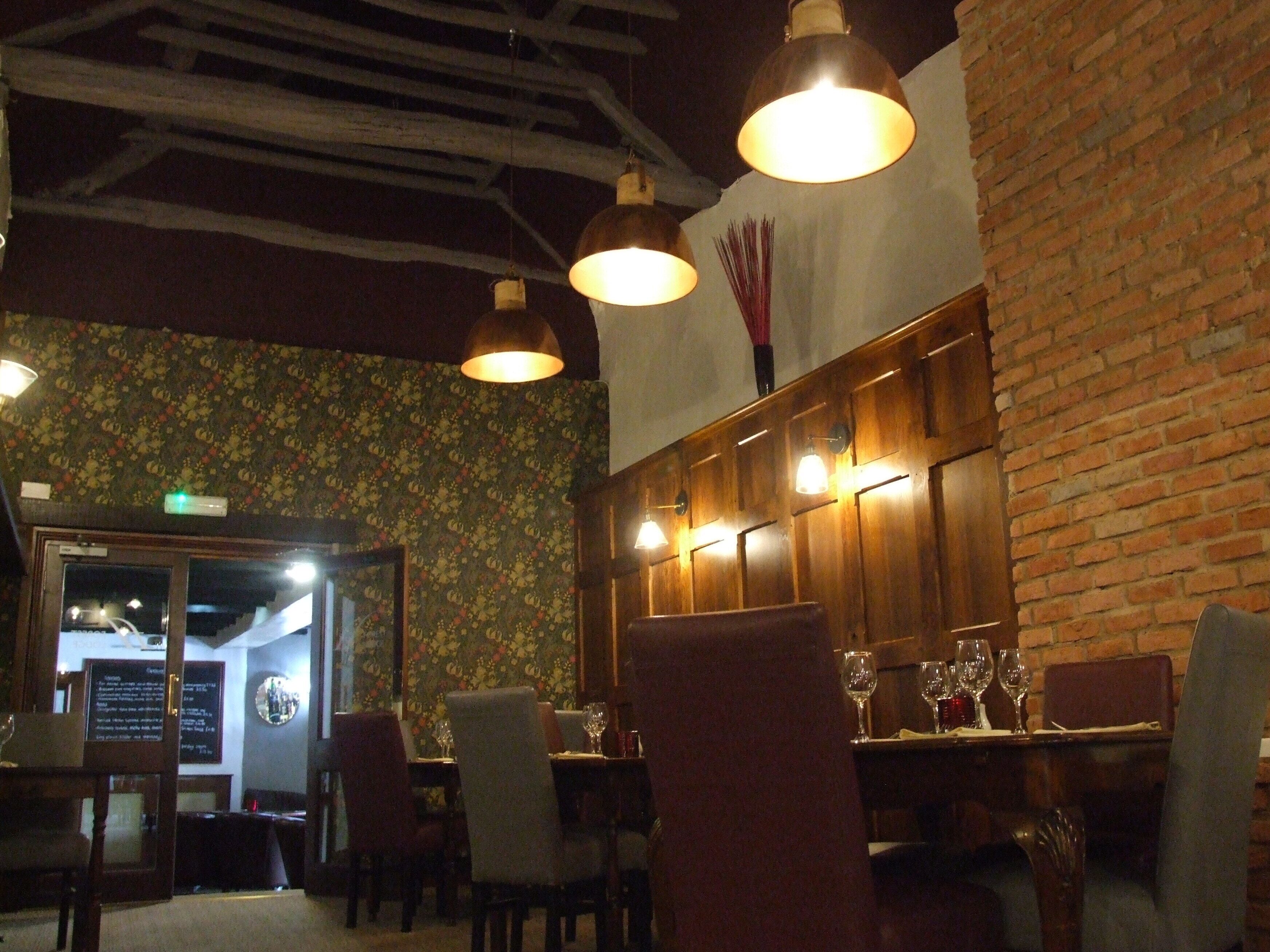 A restaurant interior with wooden paneling, hanging pendant lights, and a brick wall. Tables are set with wine glasses.