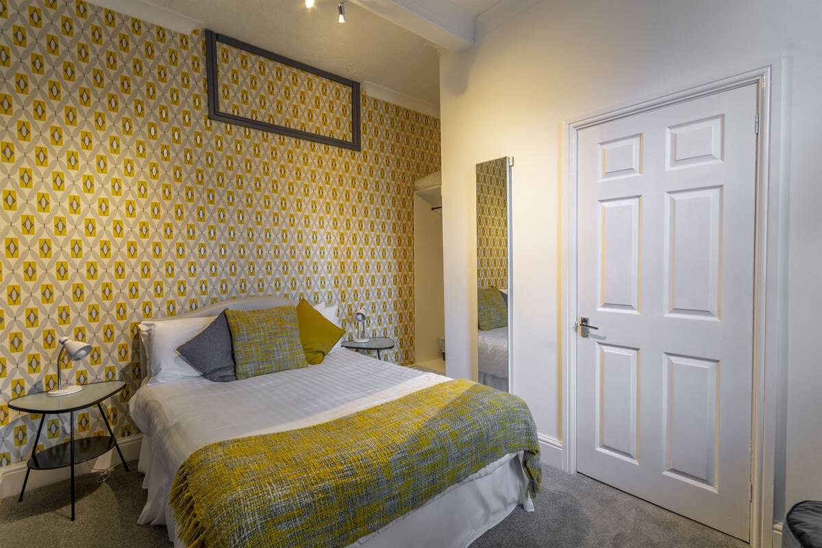 A bedroom with a patterned wallpaper, a bed with a green and white blanket, a bedside table with a lamp, and a white door.