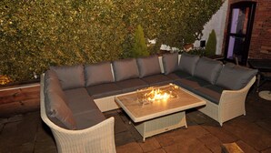 A sectional sofa with a glass-top coffee table and a fire feature, set against a brick wall with a green hedge. - The Forest Lodge (Mansfield)