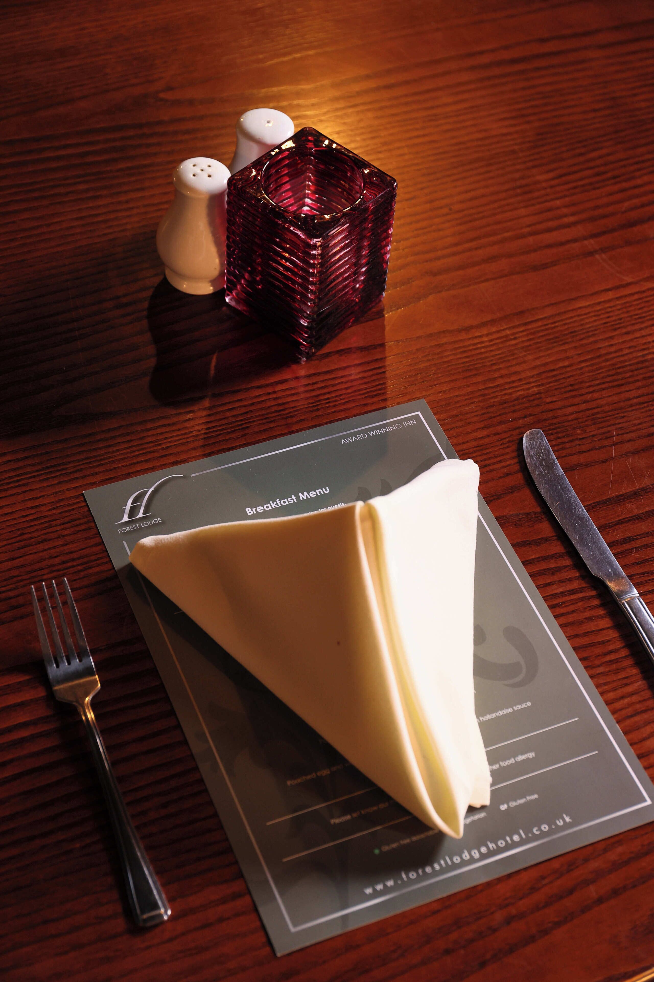 A neatly arranged breakfast setting with a menu, a folded napkin, salt and pepper shakers, a knife, and a fork on a wooden table.