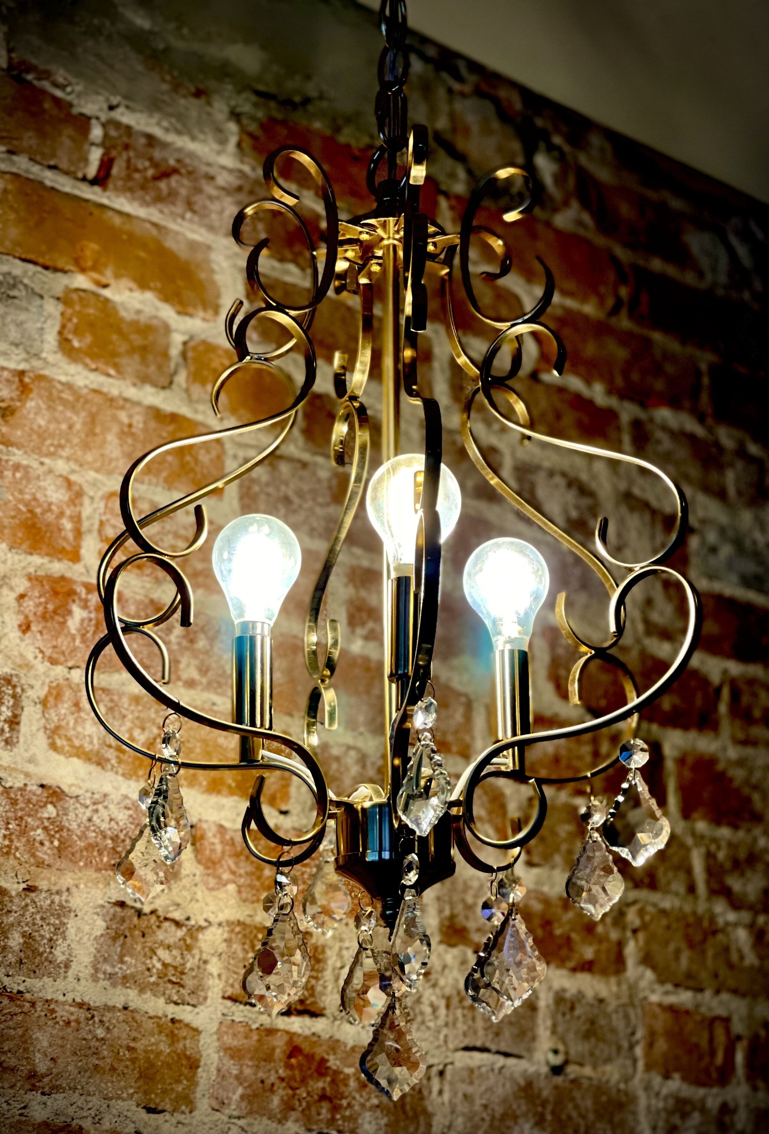 A chandelier with three light bulbs and crystal decorations against a brick wall.