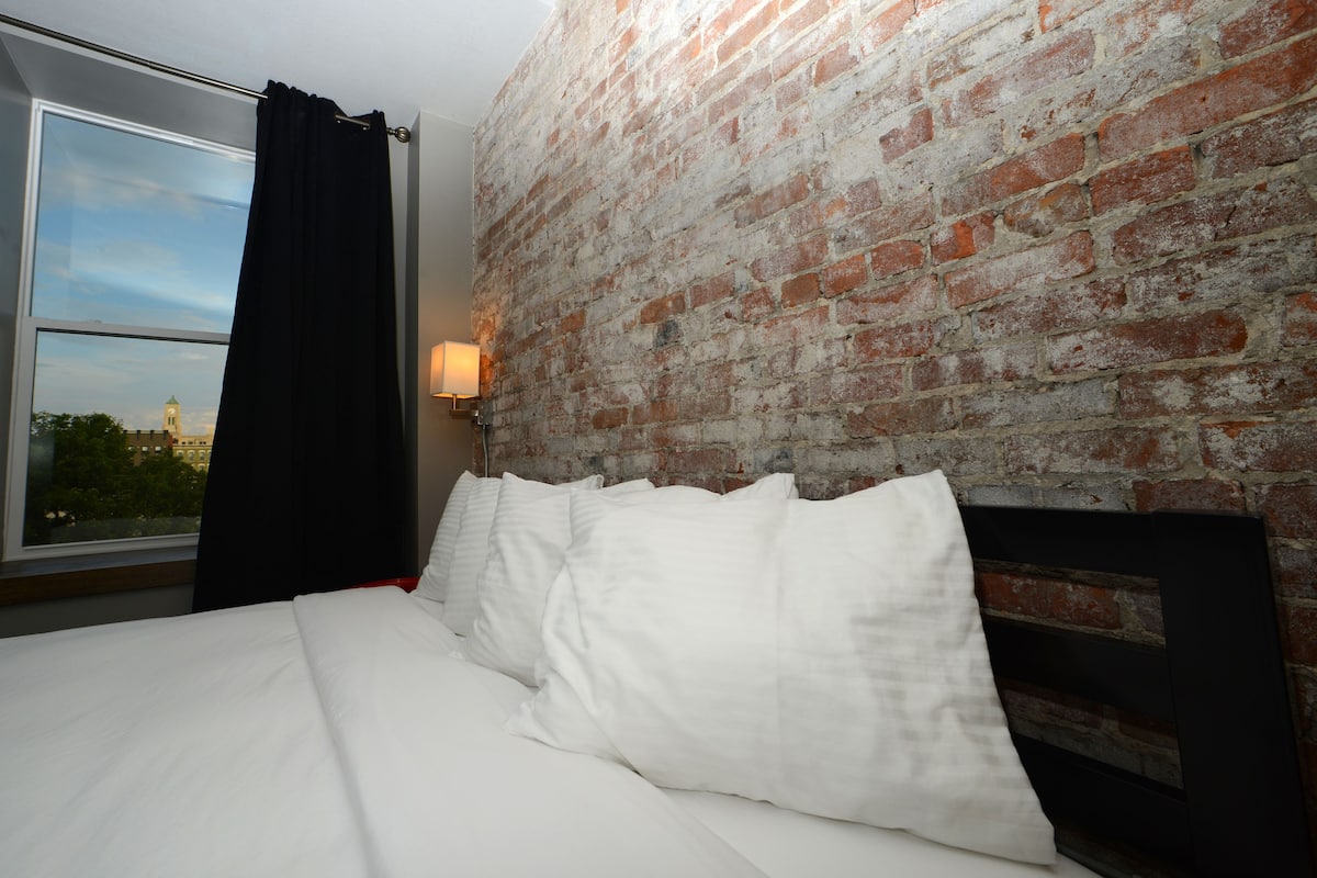 Une chambre d’hôtel avec un mur en briques, un lit avec une literie blanche, une fenêtre donnant sur un immeuble et une table de chevet avec une lampe.