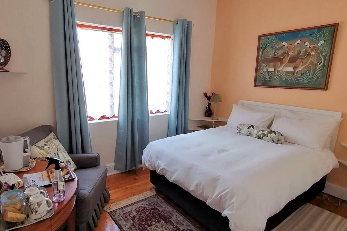 Un dormitorio ordenado con una cama grande, una mesita de noche con lámpara, una pequeña mesa con jarrón y tazas, y una ventana con cortinas.