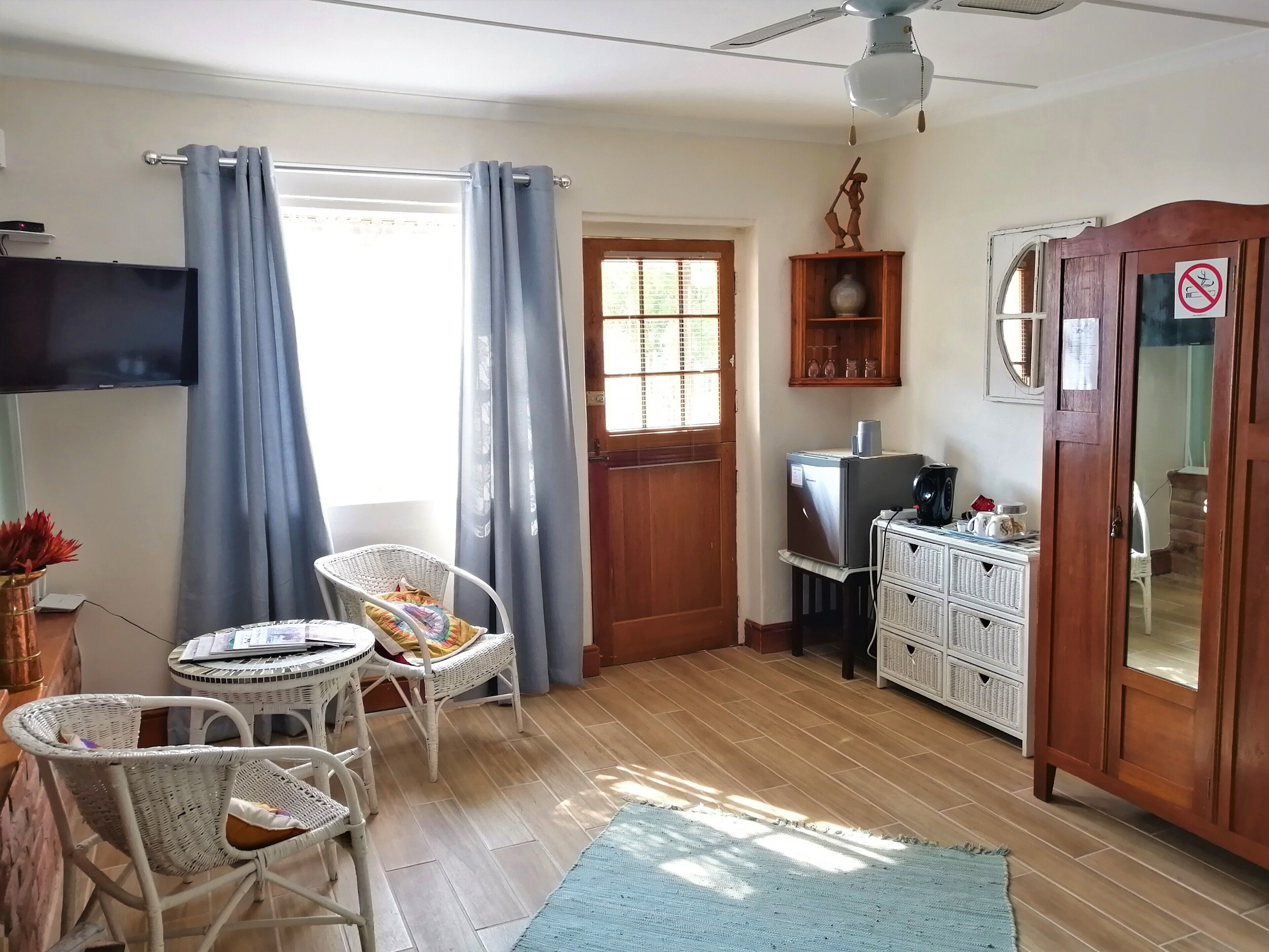 Una habitación con piso de madera, ventilador de techo, televisión, refrigerador, gabinete de madera y una puerta con letrero de 'no fumar'.