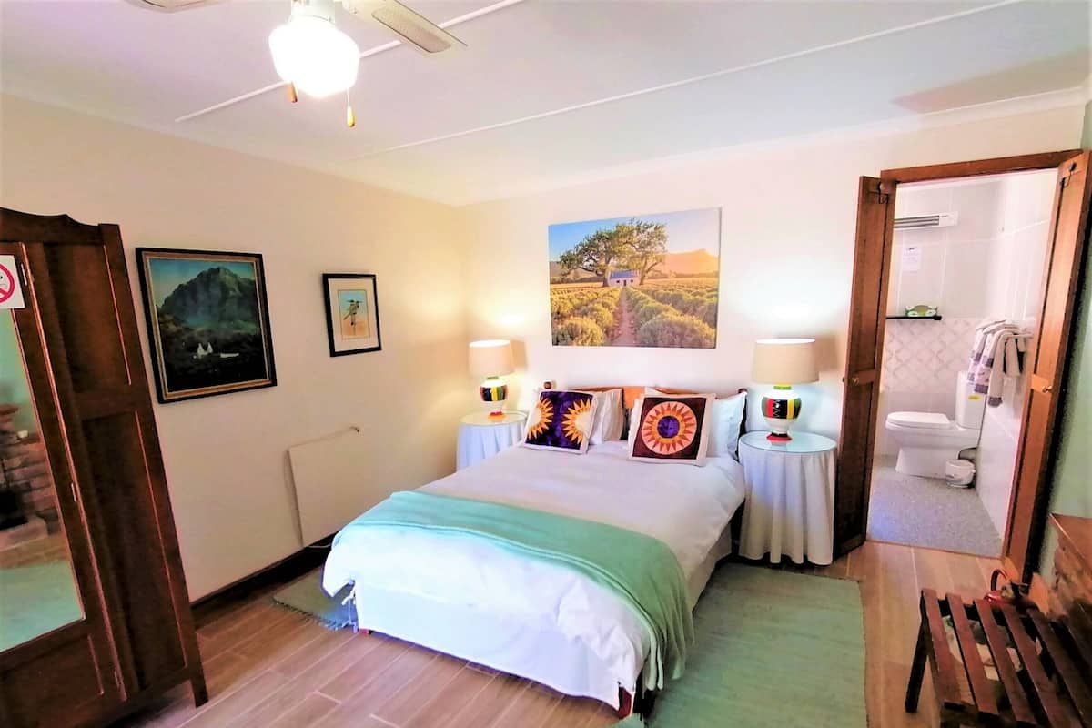 Un dormitorio con una cama, dos mesitas de noche con lámparas, un banco de madera, un baño con inodoro y una puerta que da a otra habitación.