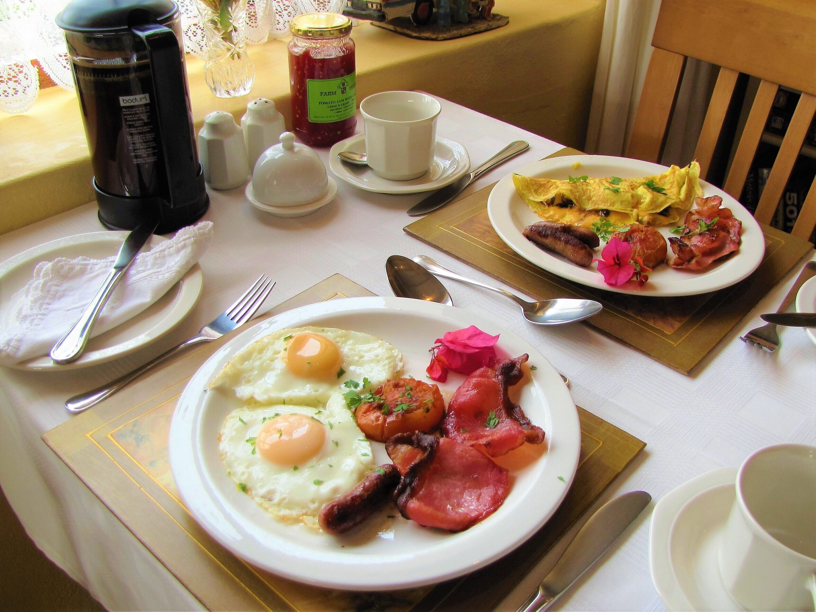 Un desayuno servido con platos de huevos, tocino y salchichas, una cafetera y condimentos sobre una mesa.