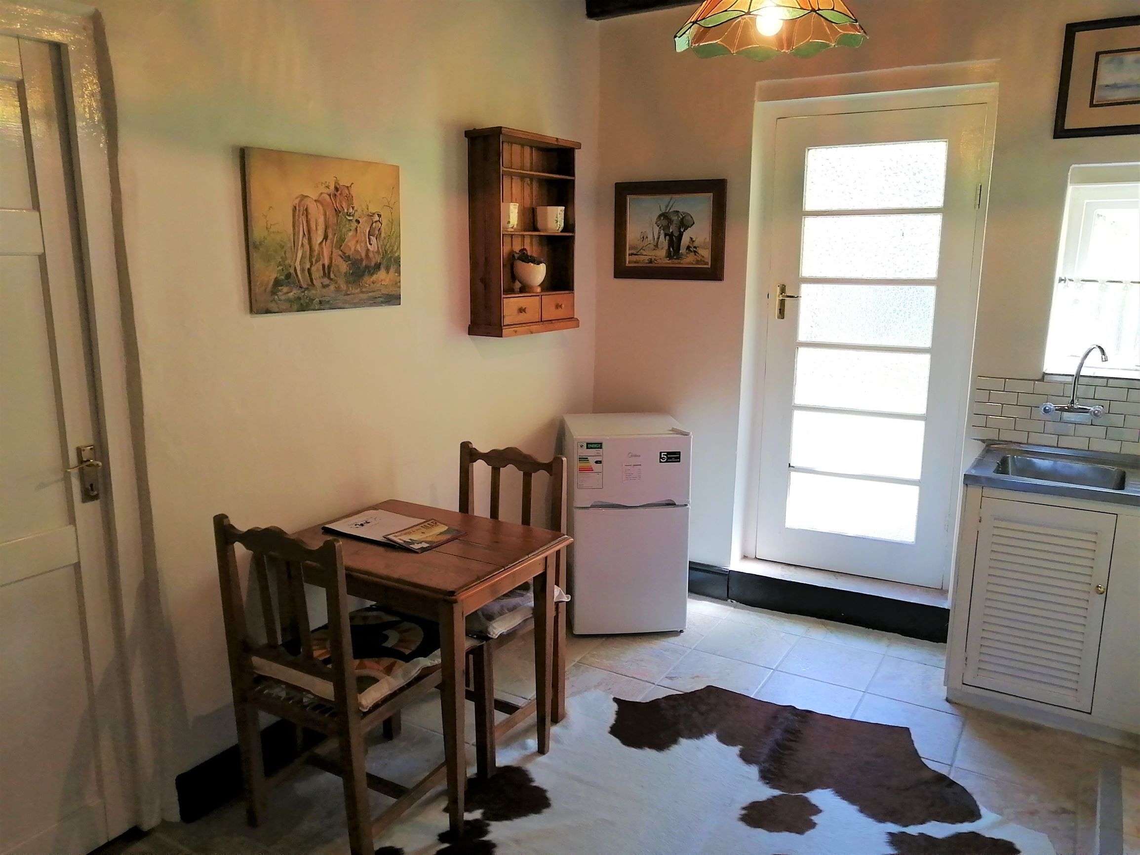 Een kleine keuken met een koelkast, een eettafel met stoelen, een kastje, en een kuitenvachten tapijt op de vloer.