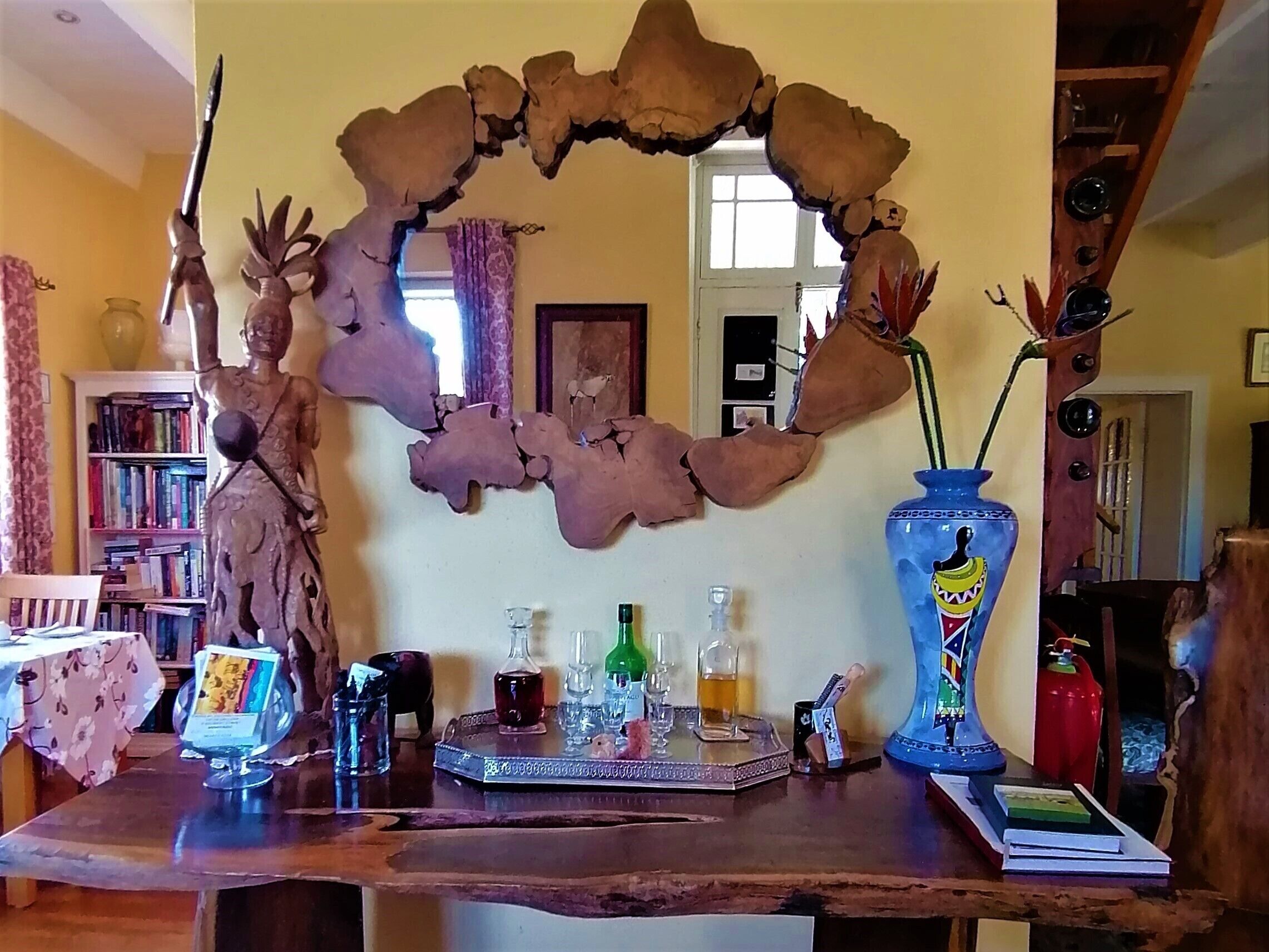 Une table en bois ornée d’une arche décorative en forme de feuilles, une statue, un vase bleu contenant une plante, ainsi que diverses bouteilles et verres.