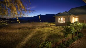 A house with a well-lit window, surrounded by a grassy yard and a small garden. - Dynasty Red Mountain Ranch (Clarens)