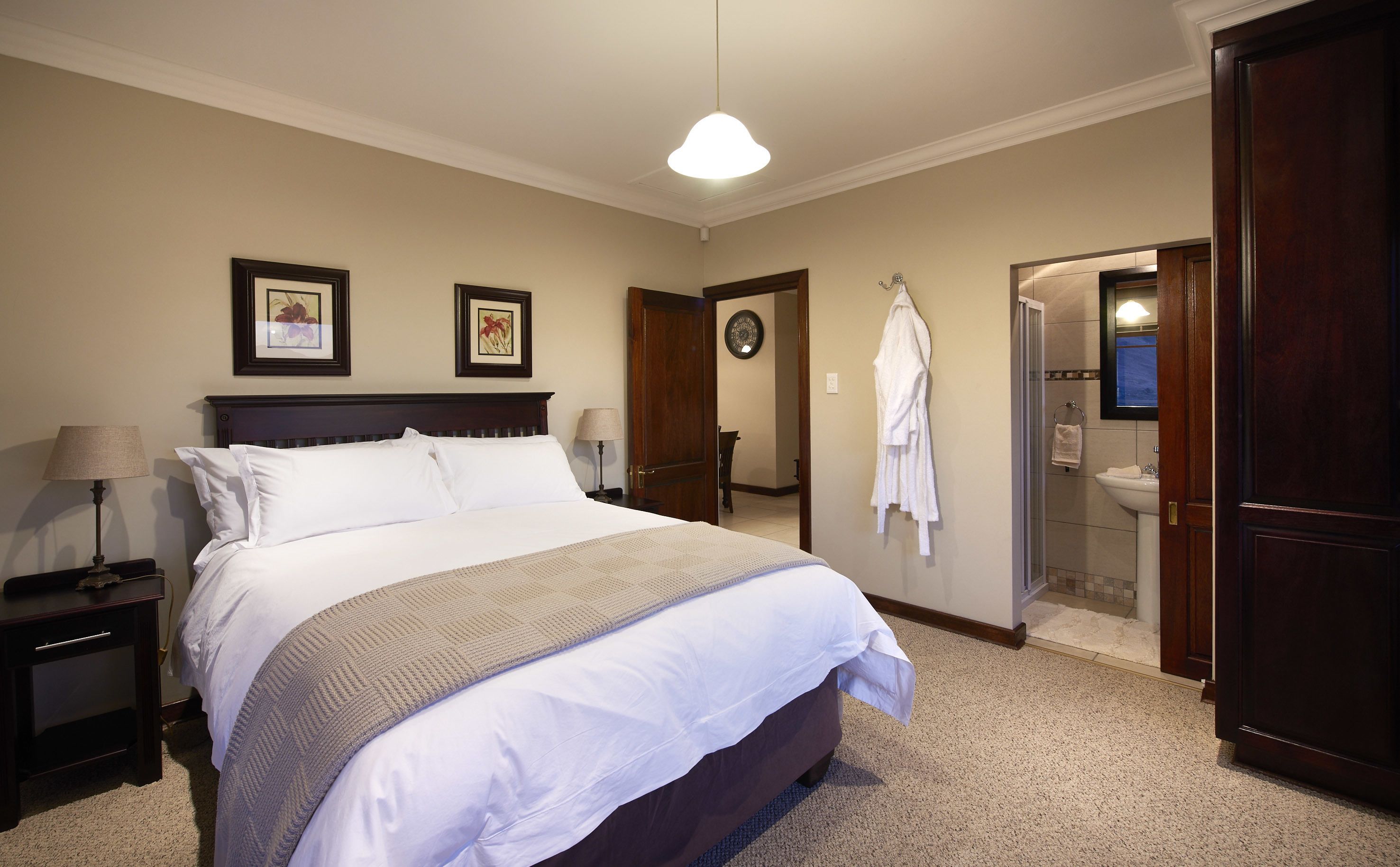 A neatly arranged bedroom with a large bed, bedside tables, a lamp, framed artwork, a door leading to a bathroom, and a robe hanging on the wall.
