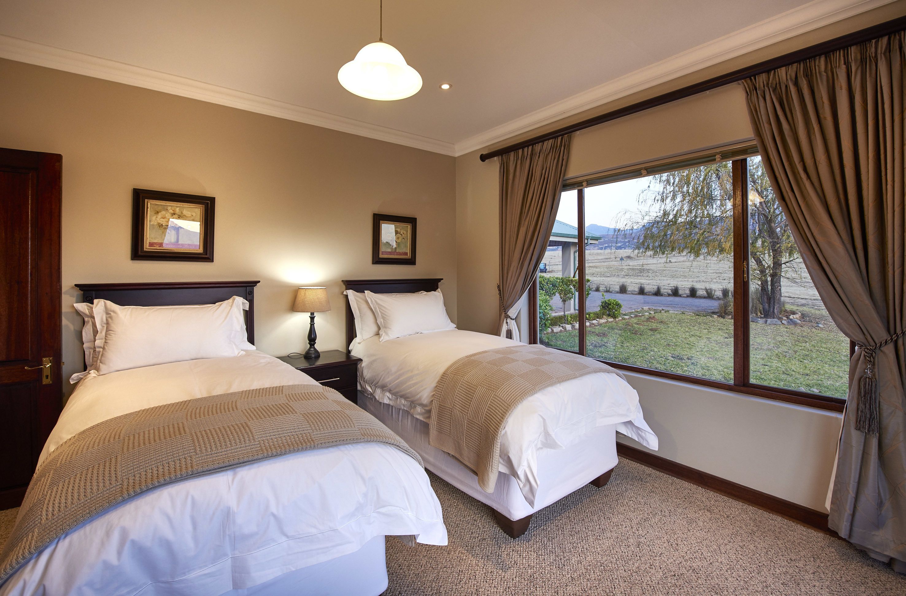 A hotel room with two beds, a large window with curtains, a lamp, and framed artwork on the wall.