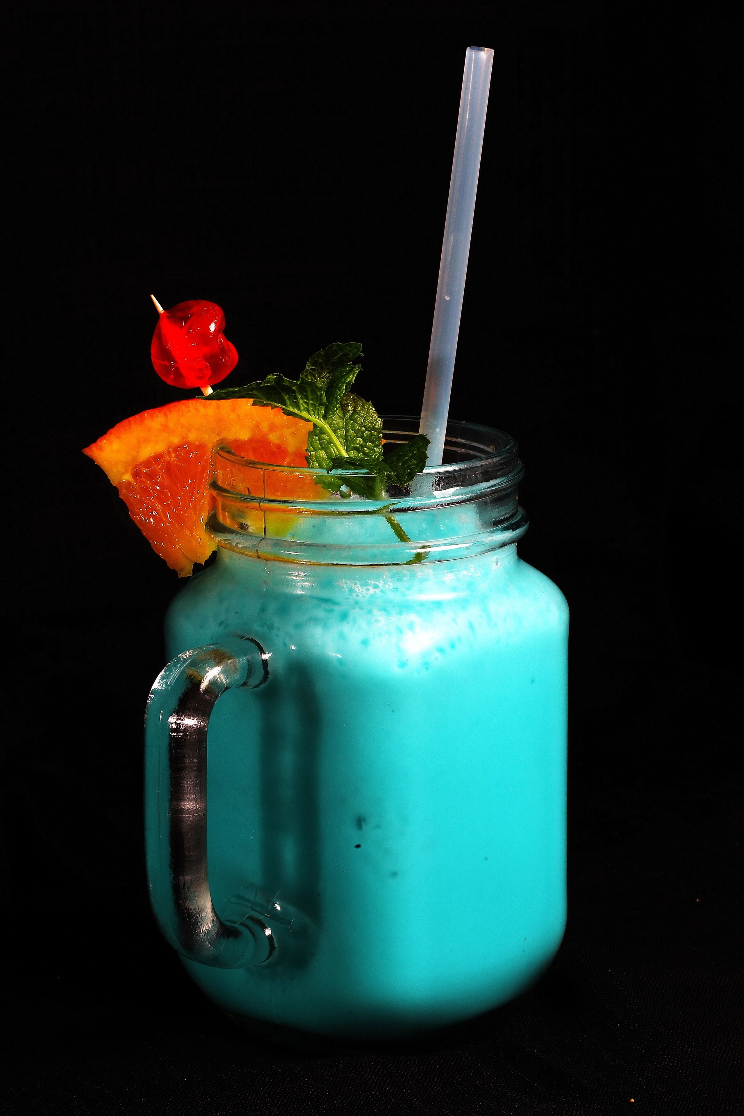 Bebida turquesa con pajita, adornada con una rodaja de naranja y una fruta roja, en un vaso de vidrio transparente.