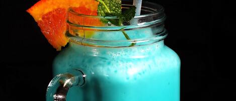 Bebida turquesa con pajita, adornada con una rodaja de naranja y una fruta roja, en un vaso de vidrio transparente.