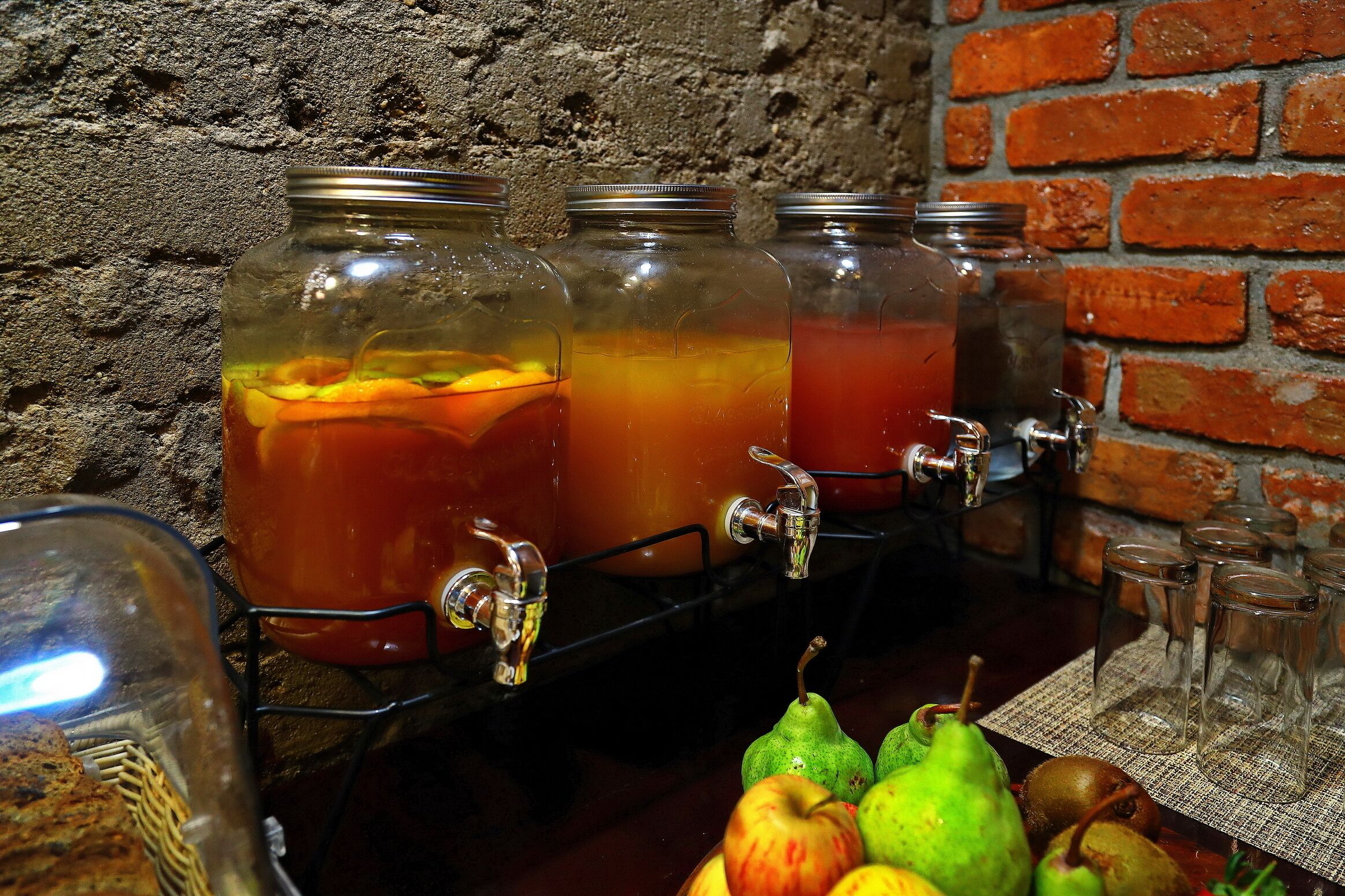 Una estación de bebidas con tres frascos de vidrio llenos de bebidas, un grifo y una mesa con frutas y vasos.