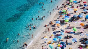 Ein überfüllter Strand mit Menschen, die schwimmen und in der Sonne liegen, unter bunten Sonnenschirmen.