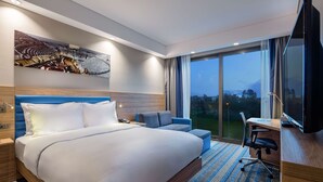 Ein Hotelzimmer mit einem großen Bett, einem Schreibtisch mit Stuhl, einem Fernseher und einem großen Fenster mit Blick ins Freie.