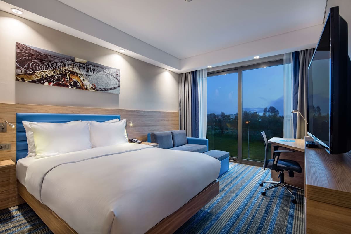 Een hotelkamer met een groot bed, een bureau met stoel, een televisie en een groot raam met uitzicht op het omliggende landschap.