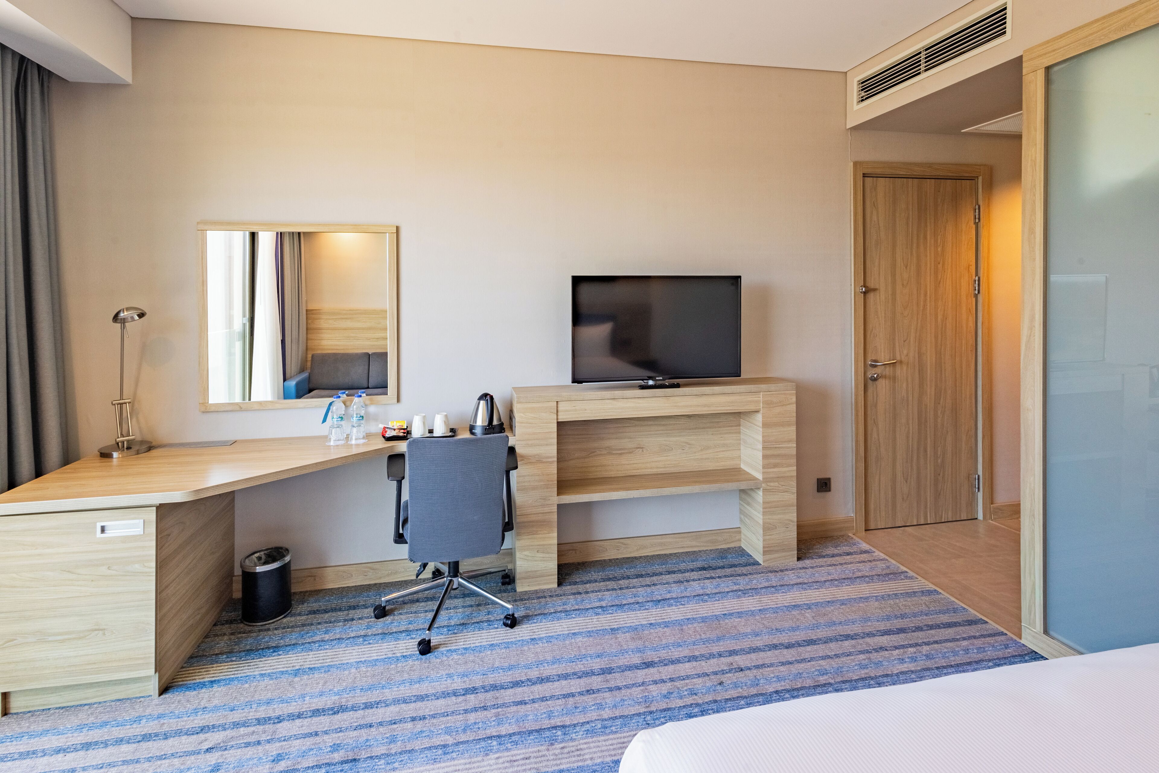 A hotel room with a wooden desk, a television, a chair, a bed, and a large window.