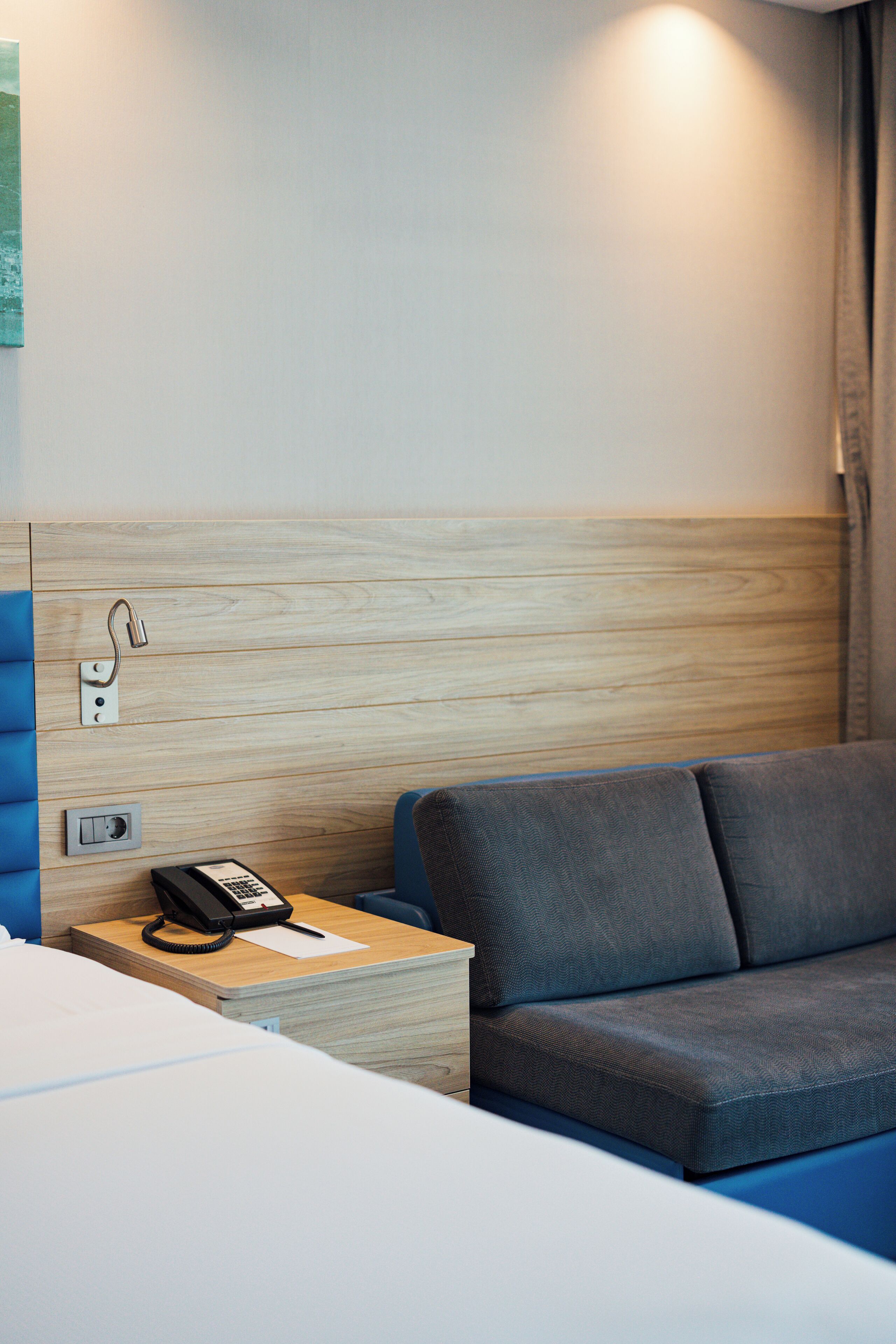 A hotel room with a wooden headboard, a grey sofa, a bedside table with a telephone, and a wall-mounted light.